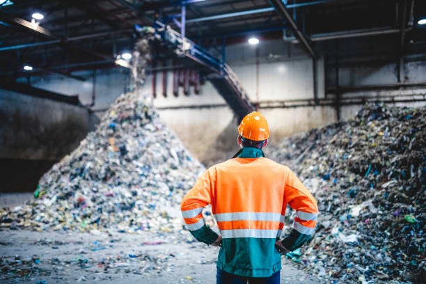 Worker in safety gear standing in front of large plastic waste pile inside recycling facility